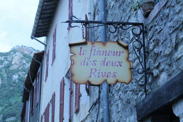 A quaint street view featuring a rustic sign hanging from a wrought iron bracket on a stone wall. The sign reads 'le fl&acirc;neur des deux Rives' in a calligraphic font. A pot of greenery is perched atop the wall. Traditional shuttered windows and a glimpse of distant rocky hills complete the serene setting.