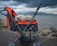 Two small white fluffy dogs are sitting comfortably in a stroller designed for pets. The stroller is predominantly black and orange and is positioned on a pathway near a body of water. The background features a cloudy sky and distant mountains, creating a serene and picturesque setting.