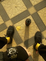 A geometric tile pattern forms the background, featuring a central grid of interlocking designs in beige and dark blue. A pair of black sneakers with bright yellow socks is visible, along with a brown water bottle standing upright. A single yellow leaf lies on the patterned tiles, and a hand wearing a black watch is seen in the foreground.