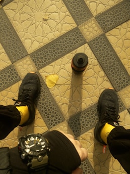 A geometric tile pattern forms the background, featuring a central grid of interlocking designs in beige and dark blue. A pair of black sneakers with bright yellow socks is visible, along with a brown water bottle standing upright. A single yellow leaf lies on the patterned tiles, and a hand wearing a black watch is seen in the foreground.