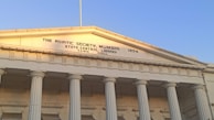 A neoclassical building with tall, white columns supporting a triangular pediment. The sun casts a warm glow on the building's facade, highlighting the inscription 'The Asiatic Society, Mumbai 1804 State Central Library Town Hall'. The sky above is clear and blue.