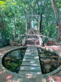 A serene outdoor setting in a lush forest with a circular stone pond at the foreground. A wooden plank walkway crosses the pond leading to stone steps and a small altar. The altar features a statue encased in glass, surrounded by vibrant green foliage, creating a peaceful and contemplative atmosphere.
