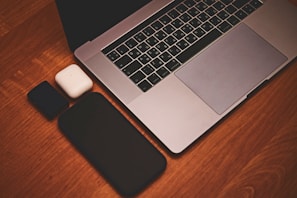 A laptop with a silver finish sits on a wooden surface. Nearby, there is a black smartphone, a white charging case, and a black rectangular power bank.