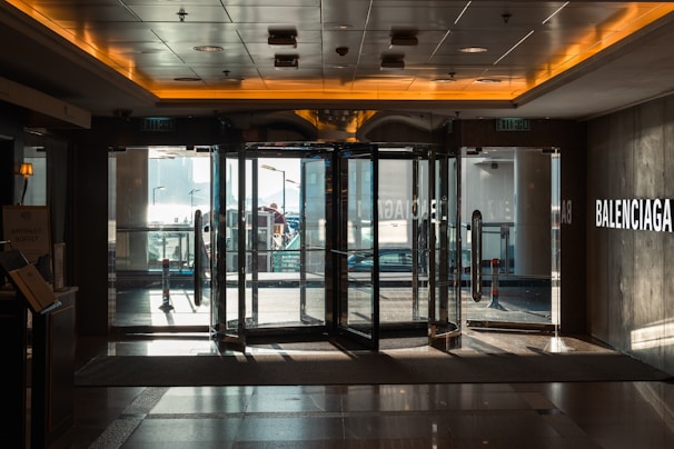 A dimly lit interior view of a building entrance featuring a revolving door, with the brand name Balenciaga visible on the wall. Sunlight streams through the glass doors, casting reflections on the polished floor. The atmosphere inside is calm, contrasting with the bright view of the outdoor area seen through the glass.