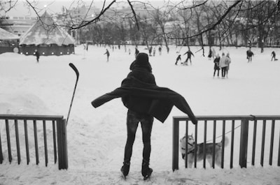 A person is seen standing with arms outstretched, wearing ice skates, and facing an outdoor ice rink filled with people skating. Snow covers the ground and a small dog stands nearby, leashed to a wooden fence. In the background, trees are bare and a building with twinkling lights is visible.
