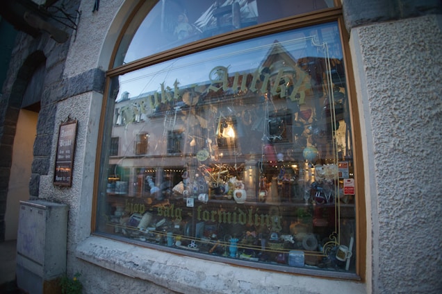 A shop window display featuring a variety of colorful objects and trinkets, including ceramics, glassware, and plush toys. The large glass window shows reflections of surrounding buildings and has gold lettering across it. The exterior of the building is made of stone and stucco with a wooden frame around the window.