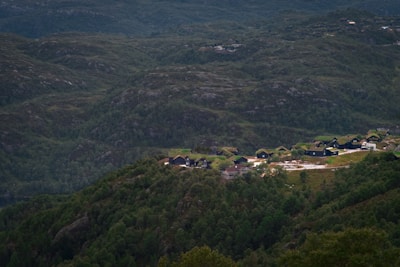 A small cluster of dark-colored houses with green roofs is nestled among lush, verdant hills. The surrounding landscape consists of rolling forests and rugged terrain, creating a peaceful, secluded environment.