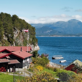 A scenic coastal landscape featuring a cluster of wooden buildings with red roofs nestled by the water. A Norwegian flag flutters on a pole, surrounded by lush greenery. The clear blue sea stretches out towards a mountainous horizon under a partly cloudy sky.