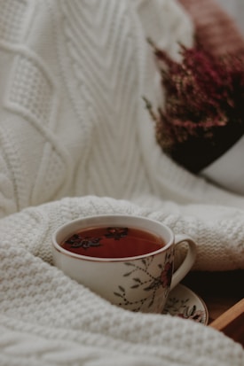 A floral-patterned teacup filled with tea is nestled within a knitted, cream-colored blanket. In the background, there are blurred plant elements adding cozy textures to the scene.