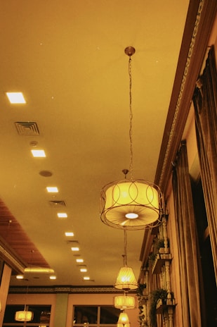 A warmly lit interior space with ornate hanging light fixtures and a series of recessed lights on the ceiling. Elegant curtains drape from the windows, and decorative plants are arranged on shelves against the walls.