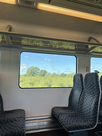 A train interior with patterned seats is visible in the foreground. Large windows provide a view of a lush, green landscape with trees under a blue sky with scattered clouds.