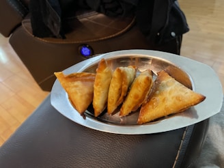 A stainless steel tray contains a row of golden-brown, crispy-fried triangular pastries placed on a dark, cushioned surface. The background features a wooden floor and part of a plush, brown upholstered furniture piece with a visible button.