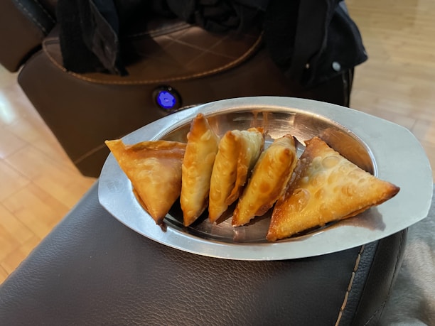 A stainless steel tray contains a row of golden-brown, crispy-fried triangular pastries placed on a dark, cushioned surface. The background features a wooden floor and part of a plush, brown upholstered furniture piece with a visible button.