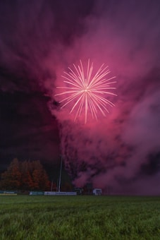 A vibrant firework explodes in the night sky, casting a pink hue over a field below. Dense smoke lingers in the air, enveloping the scene. The area is bordered by trees, and a structure with advertising hoardings can be seen in the background.