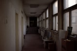 A dimly lit, narrow corridor features multiple windows on the right, some of which are foggy or dirty. The left side wall has a few doors and a fire extinguisher mounted nearby. At the far end of the hallway, there are a few steps leading to another section of the building. Various objects, including a cardboard box and stacked panels, are placed along the walls.