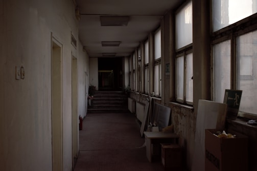 A dimly lit, narrow corridor features multiple windows on the right, some of which are foggy or dirty. The left side wall has a few doors and a fire extinguisher mounted nearby. At the far end of the hallway, there are a few steps leading to another section of the building. Various objects, including a cardboard box and stacked panels, are placed along the walls.
