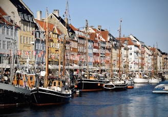 A picturesque waterfront scene featuring a row of colorful historic buildings lining a canal. Several boats are docked along the waterway, with masts and ropes visible, suggesting a bustling maritime environment. Danish flags are prominently displayed on the buildings and boats, indicating a setting in Denmark. The weather appears clear and sunny, and the perspective captures the architectural charm of the area.