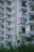 A multi-story residential building featuring numerous balconies, some with visible plants and decorations. The facade is light-colored, and the structure is characterized by its geometric form. Greenery in the foreground suggests a landscaped area nearby.