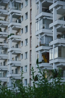 A multi-story residential building featuring numerous balconies, some with visible plants and decorations. The facade is light-colored, and the structure is characterized by its geometric form. Greenery in the foreground suggests a landscaped area nearby.