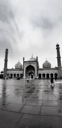 A large mosque with three prominent domes and two tall minarets, situated in an open courtyard with people walking around. The sky is overcast, enhancing the solemn and serene ambiance.