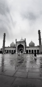 A large mosque with three prominent domes and two tall minarets, situated in an open courtyard with people walking around. The sky is overcast, enhancing the solemn and serene ambiance.