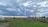 A rural landscape features a grassy field with an old, rusty piece of farm equipment on the left and a small wooden shed near the center. Cornfields line the right side, and a single tree stands in the middle background. The sky is filled with dramatic, thick clouds casting shadows on the land.