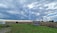 A rural landscape features a grassy field with an old, rusty piece of farm equipment on the left and a small wooden shed near the center. Cornfields line the right side, and a single tree stands in the middle background. The sky is filled with dramatic, thick clouds casting shadows on the land.