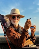An elderly man with a white beard is smiling while seated in an outdoor chair. He is wearing a wide-brimmed hat, sunglasses, and a brown suede jacket over a mustard-colored sweater. A patterned scarf is draped around his neck. In his hand, he holds a small drink glass and a smartphone.