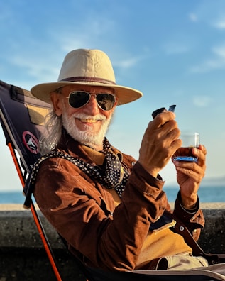 An elderly man with a white beard is smiling while seated in an outdoor chair. He is wearing a wide-brimmed hat, sunglasses, and a brown suede jacket over a mustard-colored sweater. A patterned scarf is draped around his neck. In his hand, he holds a small drink glass and a smartphone.
