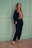 A woman stands confidently against a pastel-colored wall with white trim. She is wearing a dark suit with a pink ruffled top and black high-heeled sandals. Her long hair falls over her shoulders.