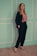 A woman stands confidently against a pastel-colored wall with white trim. She is wearing a dark suit with a pink ruffled top and black high-heeled sandals. Her long hair falls over her shoulders.
