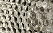 A close-up view of a textured surface with a repeating pattern resembling hexagonal cells, giving an appearance similar to a honeycomb. The material appears to be a transparent or semi-transparent plastic with subtle reflections and a silvery sheen.