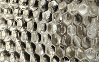 A close-up view of a textured surface with a repeating pattern resembling hexagonal cells, giving an appearance similar to a honeycomb. The material appears to be a transparent or semi-transparent plastic with subtle reflections and a silvery sheen.