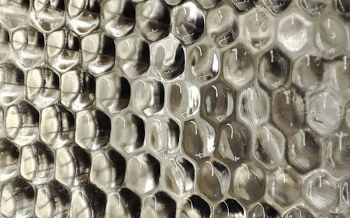 A close-up view of a textured surface with a repeating pattern resembling hexagonal cells, giving an appearance similar to a honeycomb. The material appears to be a transparent or semi-transparent plastic with subtle reflections and a silvery sheen.