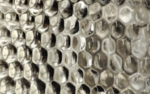 A close-up view of a textured surface with a repeating pattern resembling hexagonal cells, giving an appearance similar to a honeycomb. The material appears to be a transparent or semi-transparent plastic with subtle reflections and a silvery sheen.