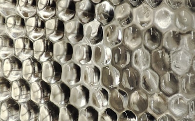 A close-up view of a textured surface with a repeating pattern resembling hexagonal cells, giving an appearance similar to a honeycomb. The material appears to be a transparent or semi-transparent plastic with subtle reflections and a silvery sheen.