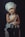 A young child wearing a chef's hat and a white apron poses with a small cooking pot and a kitchen utensil. The child has an inquisitive expression against a dark background, sitting on a cushioned stool.