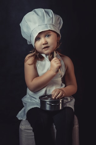 A young child wearing a chef's hat and a white apron poses with a small cooking pot and a kitchen utensil. The child has an inquisitive expression against a dark background, sitting on a cushioned stool.
