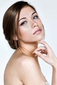 A woman with an elegant pose, showcasing smooth skin and refined features. Her hair is styled in a neat updo, and she has subtle yet sophisticated makeup highlighting her eyes and lips. The background is a simple, soft white.
