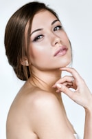 A woman with an elegant pose, showcasing smooth skin and refined features. Her hair is styled in a neat updo, and she has subtle yet sophisticated makeup highlighting her eyes and lips. The background is a simple, soft white.