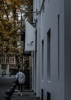 A person walks down a narrow alley lined with tall buildings. The alley is shaded by trees, and there is a sign for a boutique hotel attached to one of the buildings. The scene has a tranquil ambiance, with the autumn foliage adding a touch of color to the otherwise muted tones of the environment.