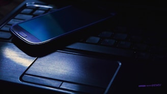 A smartphone is lying on top of a laptop keyboard in a dimly lit setting. The screen of the smartphone is off, and there is a blue hue illuminating the laptop touchpad and part of the keyboard.