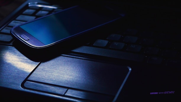 A smartphone is lying on top of a laptop keyboard in a dimly lit setting. The screen of the smartphone is off, and there is a blue hue illuminating the laptop touchpad and part of the keyboard.