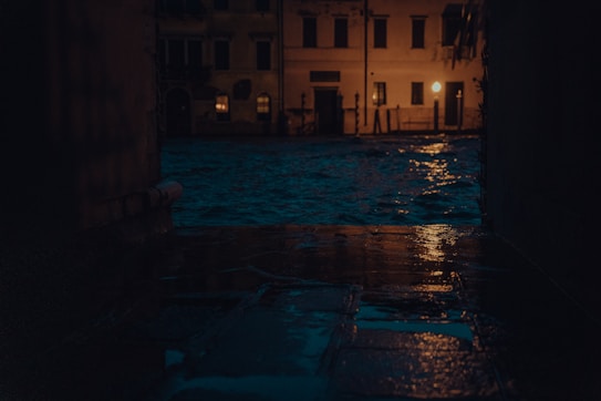 A dark, moody street scene during nighttime, showcasing the reflection of warm street lights on wet cobblestones. In the background, there are dimly lit buildings with some windows glowing from interior lights, and water appears to be overflowing onto the path.