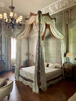 An elegant bedroom with a vintage four-poster bed adorned with ornate drapes and intricate wood carvings. The room features decorative molding on the walls and ceiling, along with a classic chandelier that casts a warm glow. The furnishings include an antique dresser and a chair, each with intricate designs, complementing the luxurious ambiance.
