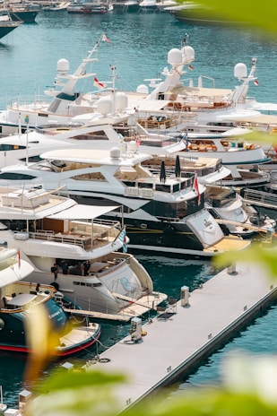 A marina filled with luxury yachts docked along the pier. The water is a clear blue and the scene is framed by vibrant green foliage. The yachts are sleek, with modern designs, and display various national flags.