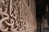 Intricate Arabic calligraphy is carved into stone, running along a wall. The letters are ornate and artistically detailed, featuring geometric and floral patterns. The image captures the texture of the stone and highlights the craftsmanship involved in creating the artwork. Soft lighting enhances the depth and shadows of the carvings.