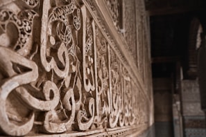 Intricate Arabic calligraphy is carved into stone, running along a wall. The letters are ornate and artistically detailed, featuring geometric and floral patterns. The image captures the texture of the stone and highlights the craftsmanship involved in creating the artwork. Soft lighting enhances the depth and shadows of the carvings.