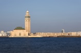 A prominent mosque stands near the waterfront, characterized by its large minaret adorned with intricate designs. The structure features a blend of traditional Islamic architecture with modern city buildings visible in the background under a clear blue sky.