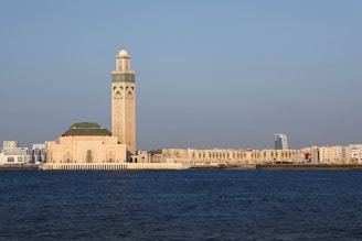 A prominent mosque stands near the waterfront, characterized by its large minaret adorned with intricate designs. The structure features a blend of traditional Islamic architecture with modern city buildings visible in the background under a clear blue sky.